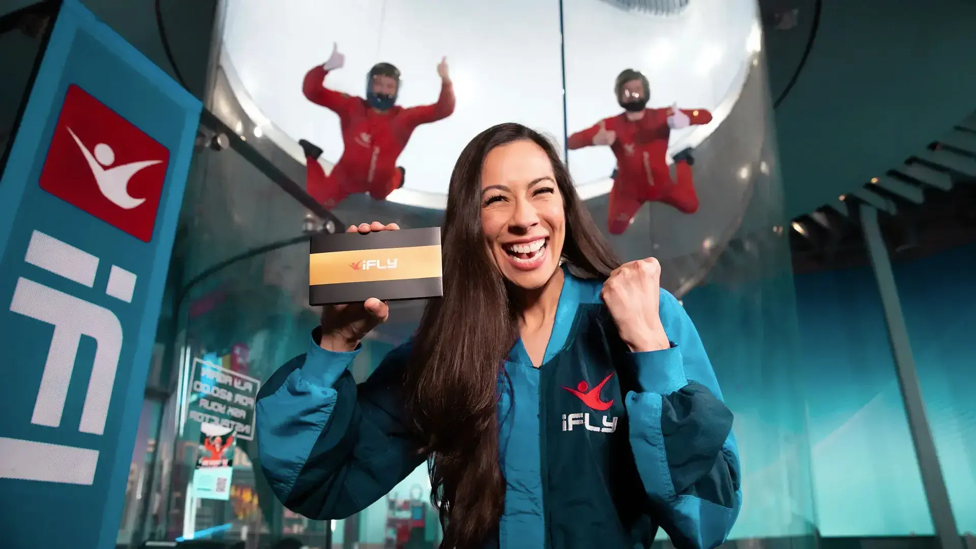 An excited woman showing off the iFLY Gift box in front of two flyers in the tunnel