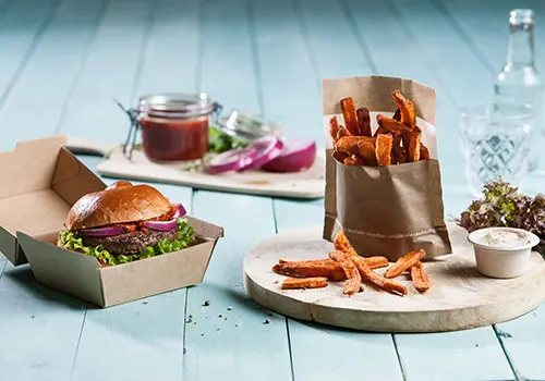 Bild eines Hamburgers in einer Pappschachtel, Süßkartoffel-Pommes in einer papiertuüte und einer kleinen Becher Dipsauce auf einem Holzbrett.