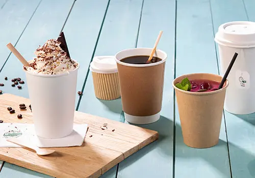 Bild von mehreren Bechern auf einer Holzoberfläche, darunter ein weißer Becher mit Sahne, ein Pappbecher und ein Becher mit Strohhalm.