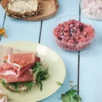 Ein Teller mit einem Sandwich, belegt mit Schinken und Grünzeug, neben Schalen mit Hackfleisch und Aufstrich auf einem blauen Tisch.