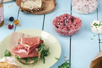 Ein Teller mit einem Sandwich, belegt mit Schinken und Grünzeug, neben Schalen mit Hackfleisch und Aufstrich auf einem blauen Tisch.