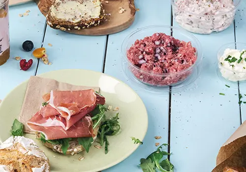 Ein Teller mit einem Sandwich, belegt mit Schinken und Grünzeug, neben Schalen mit Hackfleisch und Aufstrich auf einem blauen Tisch.