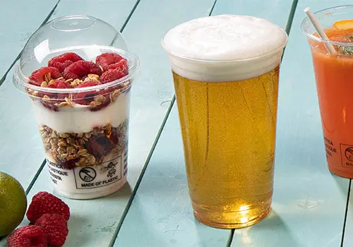 Klarer Plastikbecher mit schaumigem Bier, neben einem Parfait in einem Becher mit Himbeeren und Granola, auf einer blauen Holzfläche.