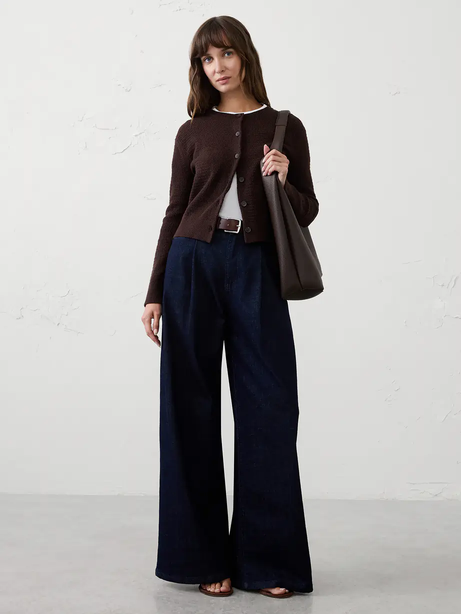 Person in a brown buttoned cardigan over a white tee, wide-leg dark denim trousers, carrying a brown shoulder bag.