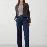 Person standing against a plain light wall wearing a dark leather jacket over a beige knit sweater, dark blue straight‑leg jeans, and black loafers.