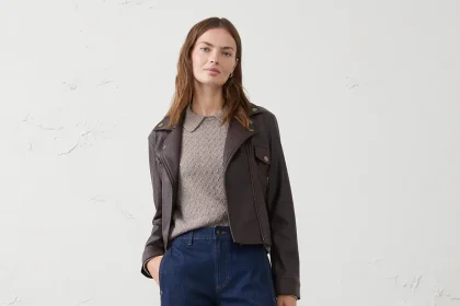 Person standing against a plain light wall wearing a dark leather jacket over a beige knit sweater, dark blue straight‑leg jeans, and black loafers.