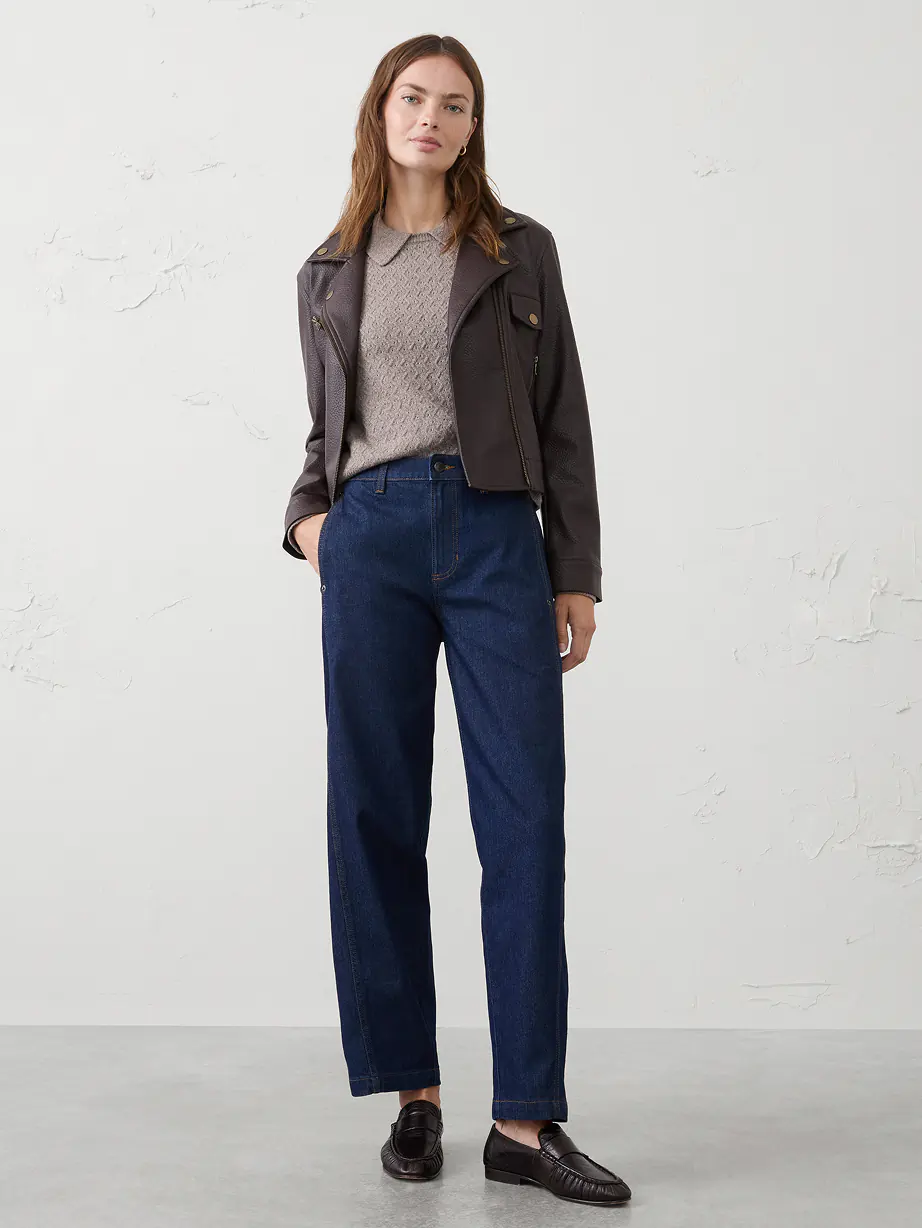 Person standing against a plain light wall wearing a dark leather jacket over a beige knit sweater, dark blue straight‑leg jeans, and black loafers.