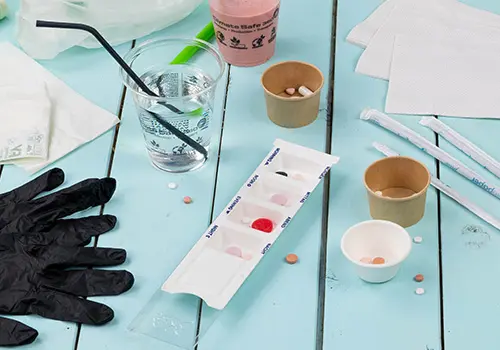 Verschiedene Gegenstände auf einem hellblauen Tisch, einschließlich eines Wasserglases, Bechern, Handschuhen und einem Medikamentenorganizer.