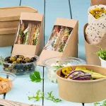 Verschiedene Lebensmittelbehälter, darunter Sandwichdosen, Salatschüsseln und Becher, auf einer hellblauen Oberfläche.