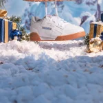 A pair of white Reebok sneakers with gum soles, suspended in a snowy winter scene with pine branches, wrapped gifts, and a blurred mountain background.