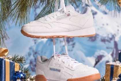 A pair of white Reebok sneakers with gum soles, suspended in a snowy winter scene with pine branches, wrapped gifts, and a blurred mountain background.