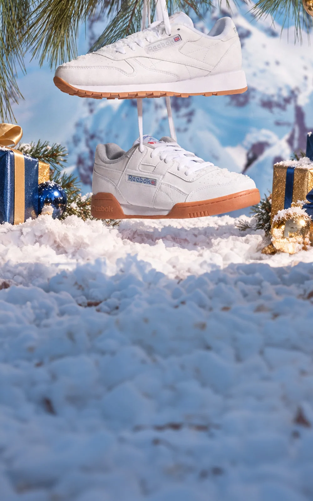 A pair of white Reebok sneakers with gum soles, suspended in a snowy winter scene with pine branches, wrapped gifts, and a blurred mountain background.